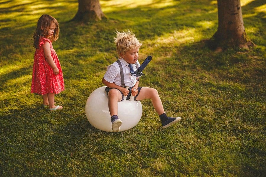 A little boy bounces on a space hopper in a field as a little girl with a red dress on watches him