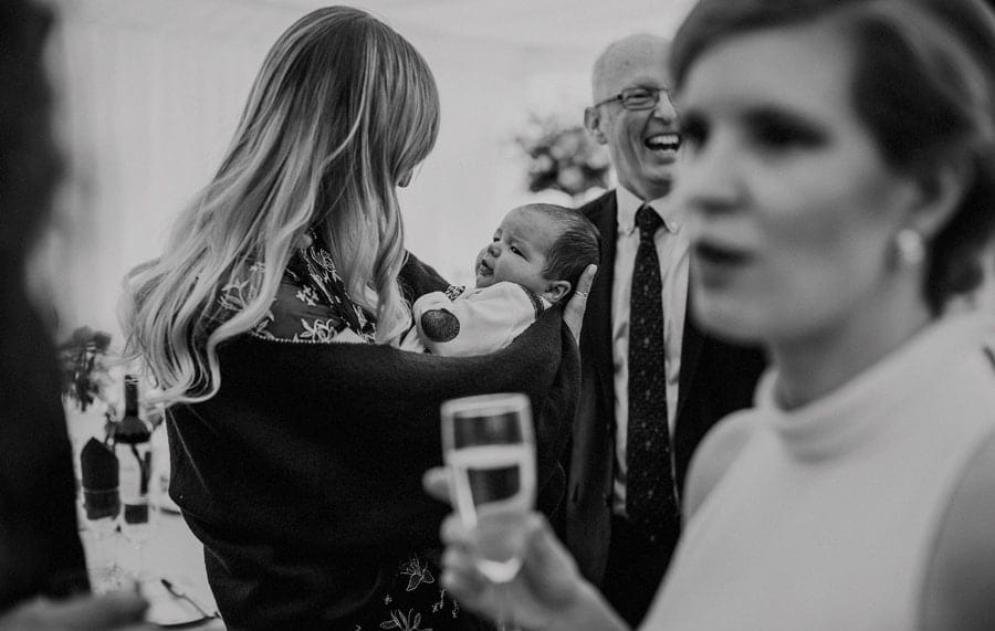 A lady holds her baby boy in her arms next to wedding guests in a marquee