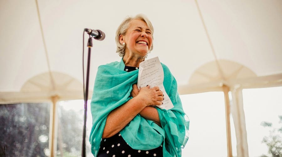 The brides mother stands in front of a microphone and smiles as she holds a piece of paper