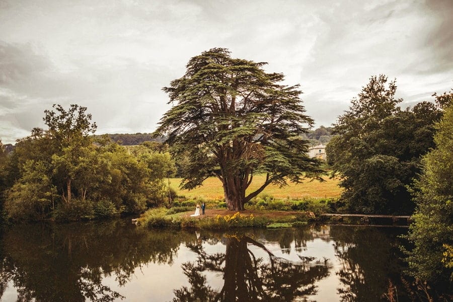 The bride and groom stand next to each other under a large tree in front a large lake