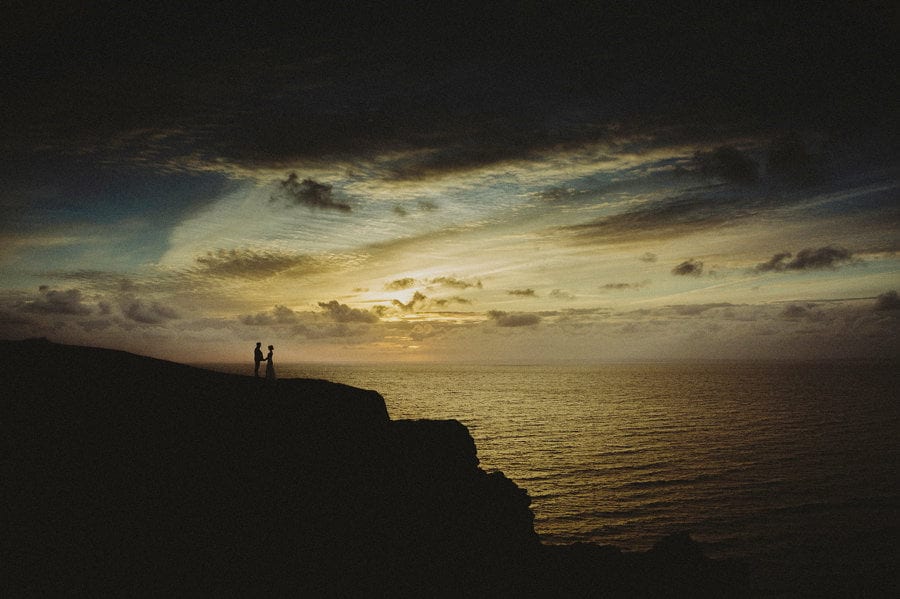 The bride and groom stand on the edge of a cliff next to the sea as the sun sets in the background