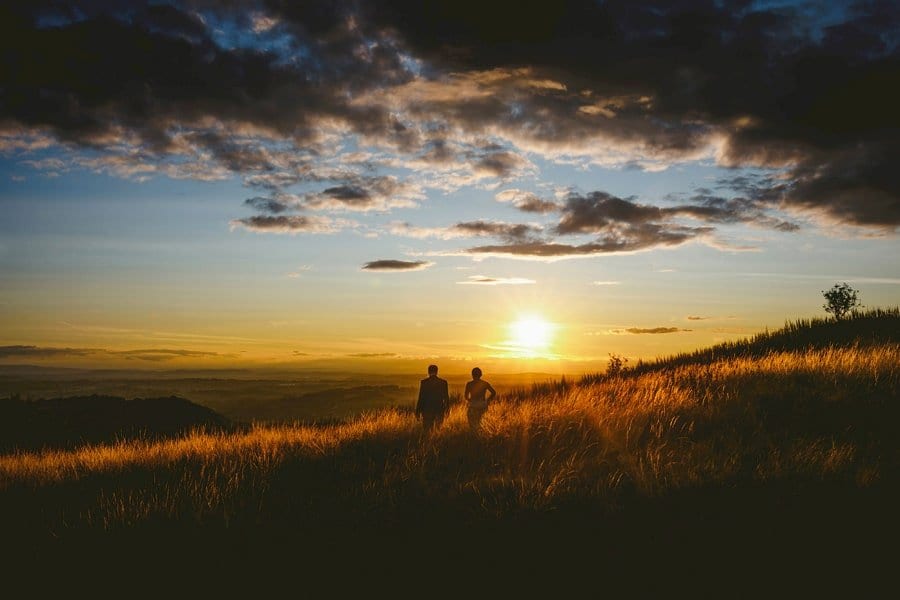 A bride and groom walk through grass next to each other on a hill as the sun goes down in the distance