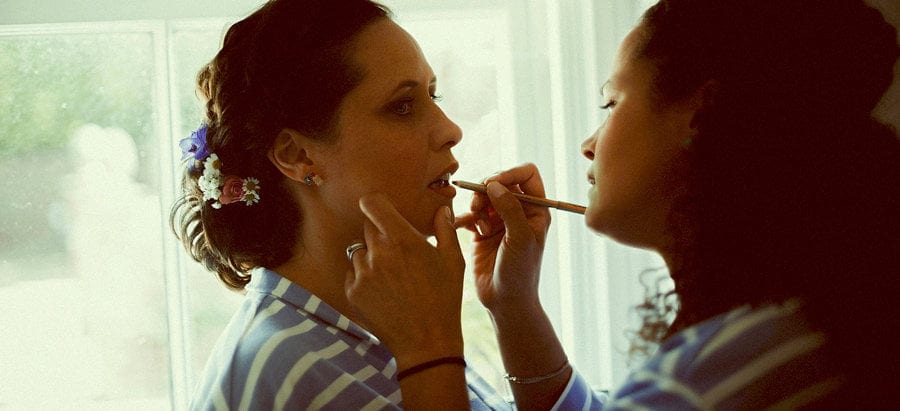 A bridesmaid applies lipstick to the brides lips next to a large window