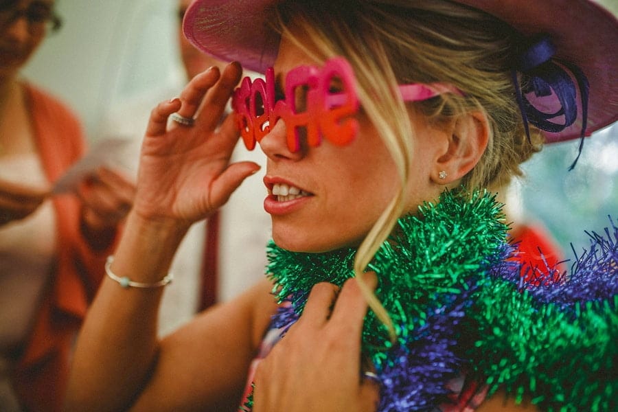 A lady wears a pink hat and holds her sunglasses in her right hand and wraps tinsel around her neck
