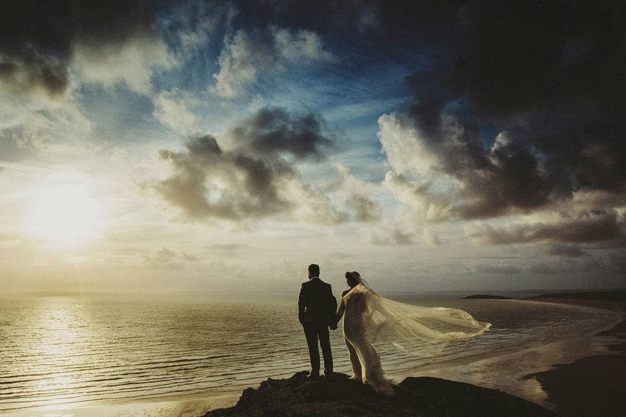 The bride and groom hold each others hand and stand next to each other on the edge of a cliff and watch the sun go down over the sea