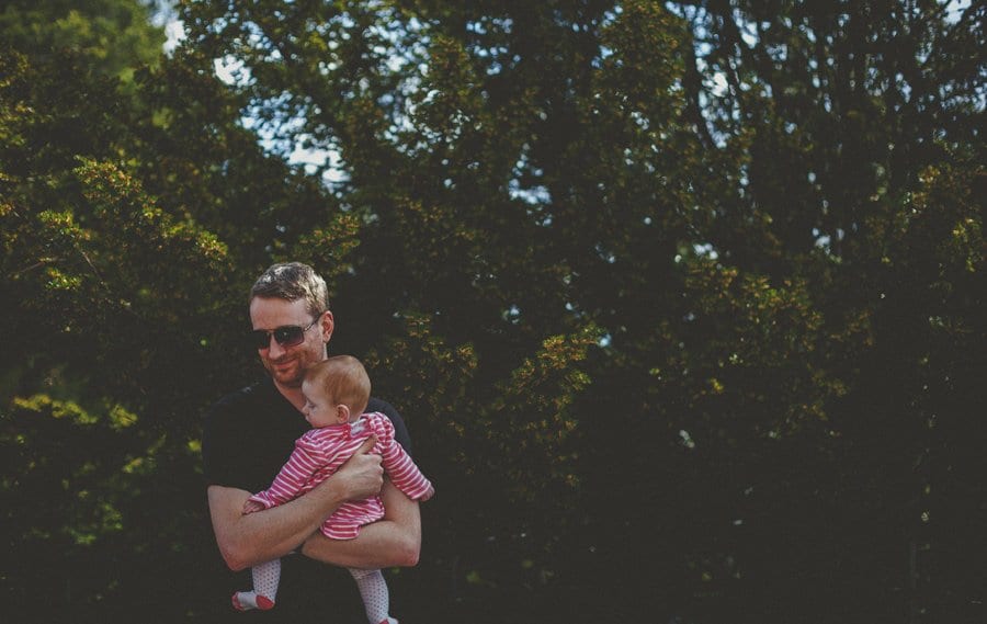 A man holds his baby daughter close to his chest in front of a large bush in the gardens at Cowley Manor Estate