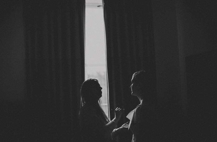 The makeup artist looks at the brides face in front of a large curtain next to a window at Cowley Manor