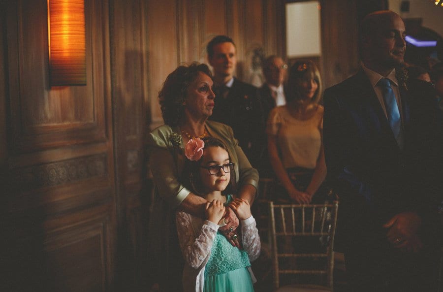 A flower girl stands and watches the wedding ceremony in front of her mother
