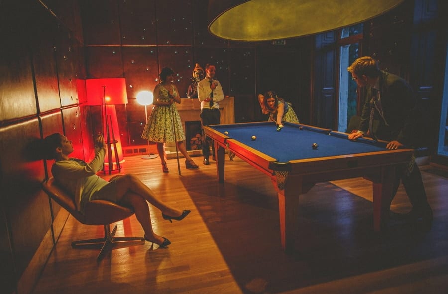A lady sat in a chair next to the pool table takes a photograph on her mobile phone of wedding guests playing pool at Cowley Manor Hotel in Cheltenham