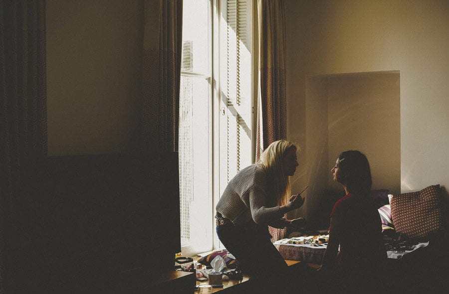 A makeup artist bends down and looks closely at the face of one of the bridesmaids
