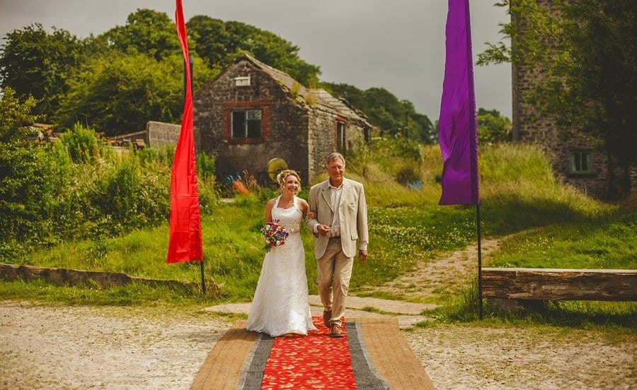 The bride holding her bouquet of flowers in her right hand and her father walk towards the outdoor wedding ceremony