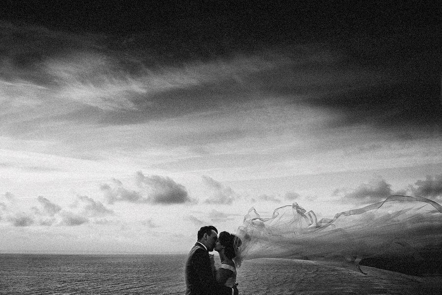 The bride and groom kiss each other in front of the sea as her veil moves in the wind