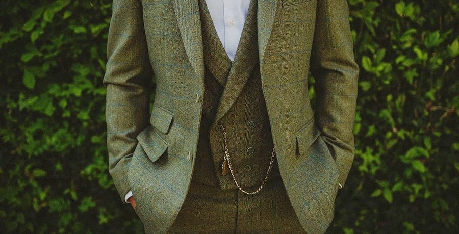 The groom stands in front of a large bush dressed in a green tweed suit with an old watch hanging from his waistcoat