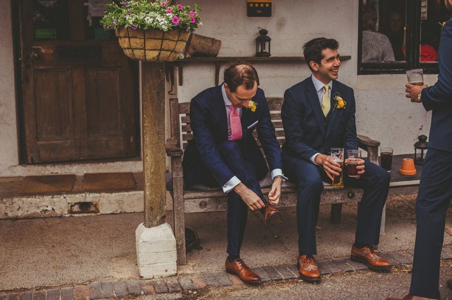 The groom sits on a wooden bench outside a public house and ties his shoelaces