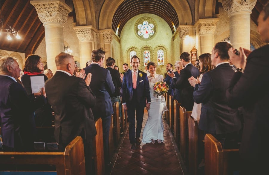 The bride and groom walk down the aisle of the church together as wedding guests stand and applaud
