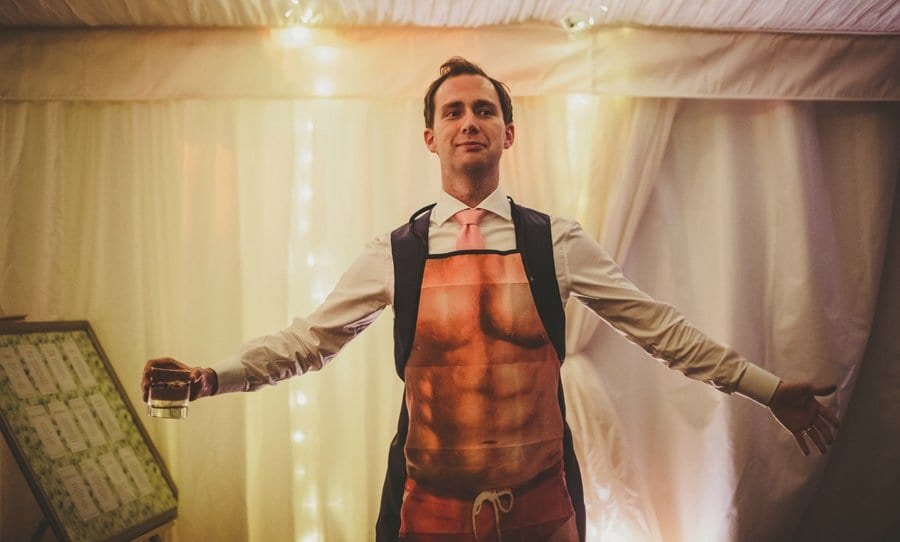 The groom stands on the edge of the dancefloor in the marquee holding a glass and wearing an apron
