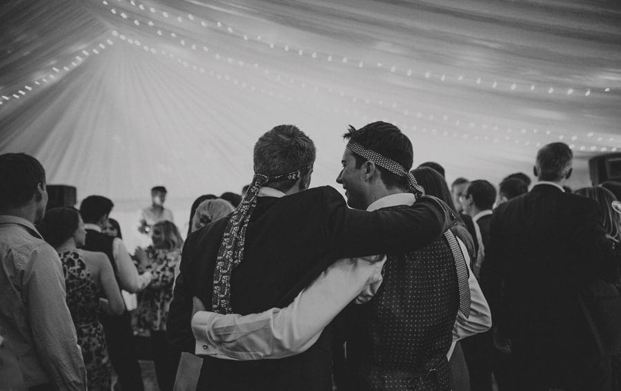 Wedding guests with ties wrapped around their heads put their arms around each other in the marquee