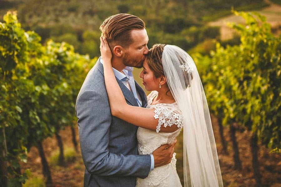 The groom holds the bride and kisses her on the forehead in a field as she puts both her hands on the back of his head