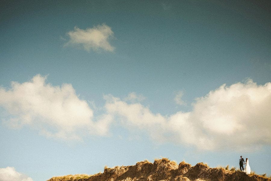 The bride and groom walk hand in hand on the top of sand dunes with blue sky and clouds above them