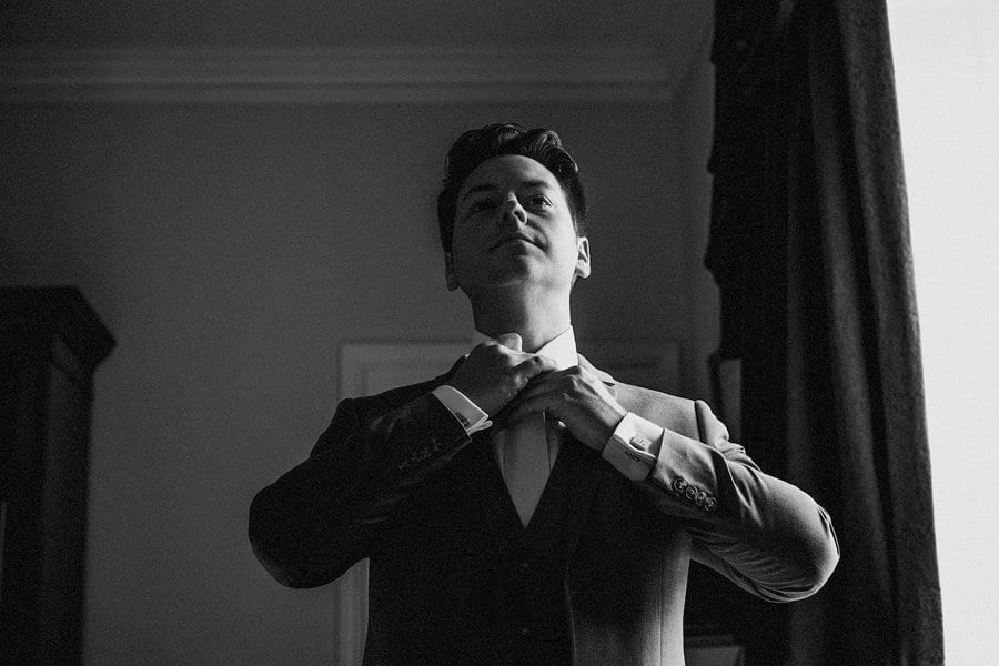 The groom looks in a mirror and straightens his tie next to a large window in the master bedroom