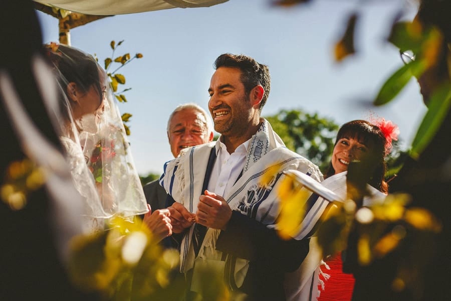 A groom stands in front of the bride and smiles as he holds a wedding ring during the outdoor wedding ceremony