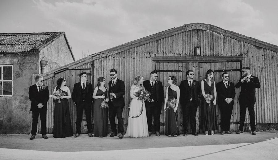 The bride and groom stand in front of large shed and talk with the wedding party