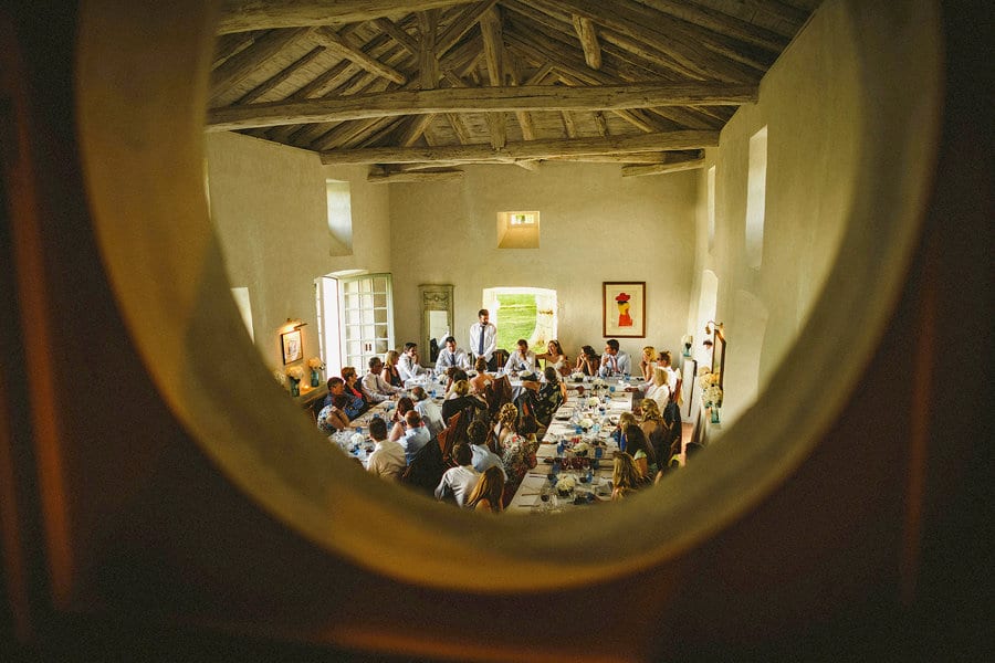 The best man stands up in front of the wedding party who are sat in front of large wooden tables and delivers his speech