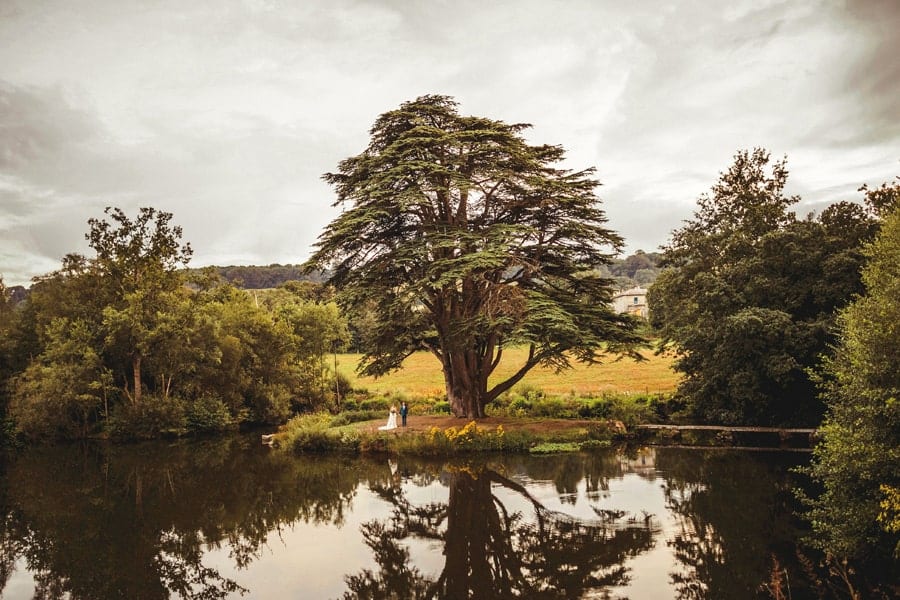 The bride and groom stand next to each other under a large tree in front a large lake