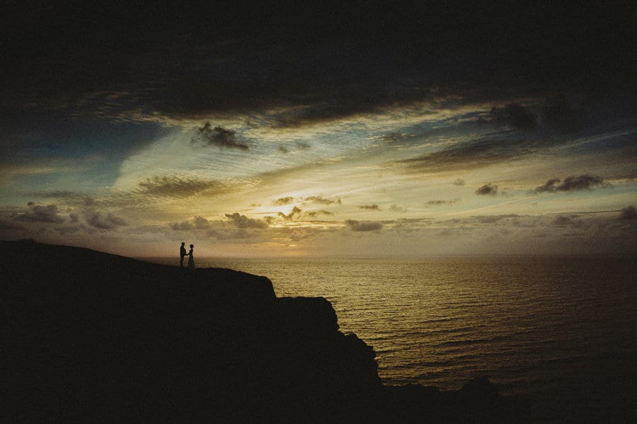 The bride and groom stand on the edge of a cliff next to the sea as the sun sets in the background