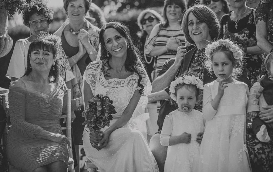 The bride sits on a chair next to her family during the outdoor wedding ceremony at Micklefield Hall