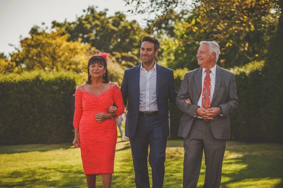 The groom and his parents walk across the garden during the outdoor wedding ceremony at Micklefield Hall