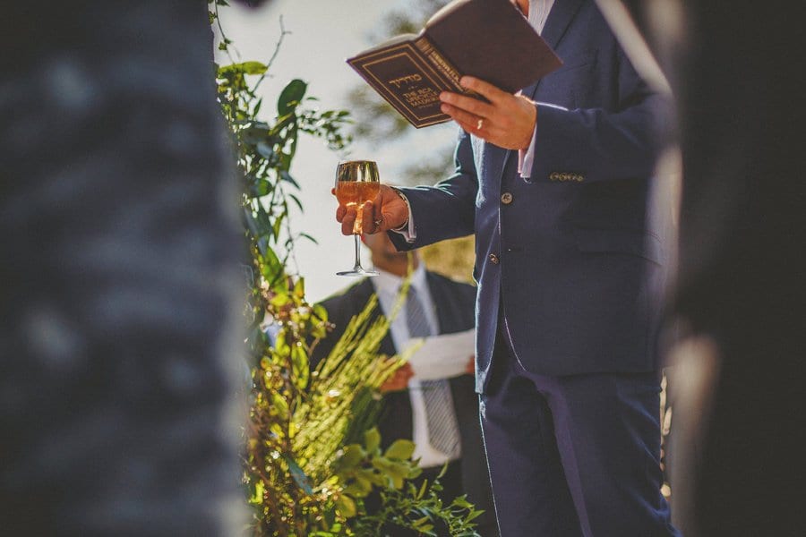 A man stands and holds a book in his left hand and a glass of wine in his right hand during the outdoor wedding ceremony at Micklefield Hall