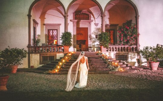 The bride walks towards stone steps as her veil is moved by the wind