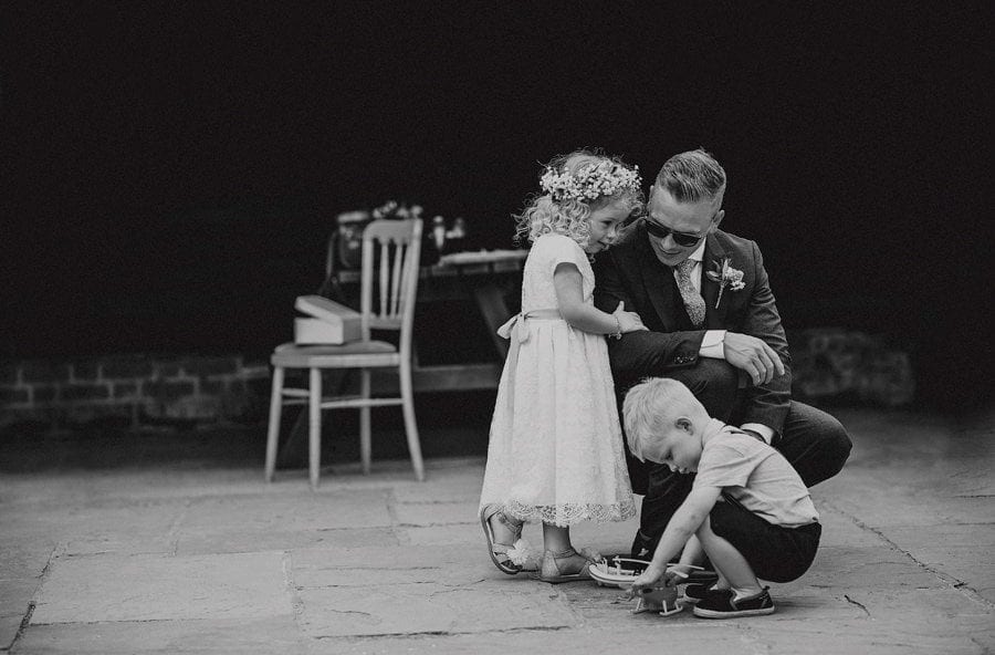 The groom kneels down and listens to his daughter in a courtyard with a little boy in front of them playing with a toy helicopter