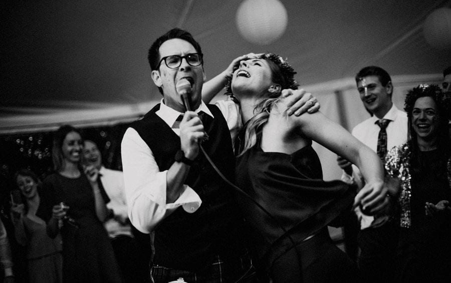 A man sings into a microphone and wraps an arm around a lady on the dancefloor of the marquee