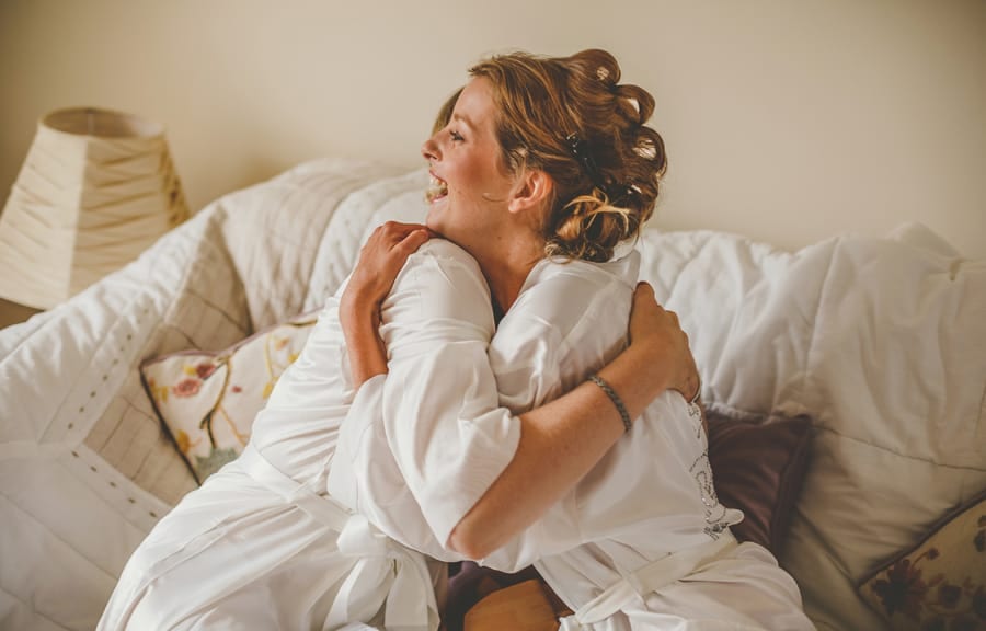The bride and her bridesmaid embrace each other as they sit on a sofa