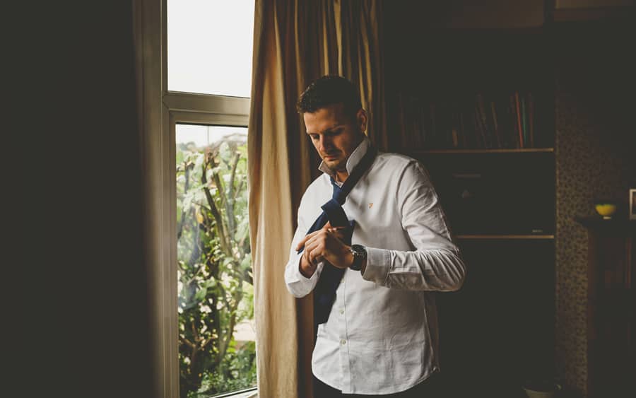 The groom stands next to a large window, looks down and puts his tie on