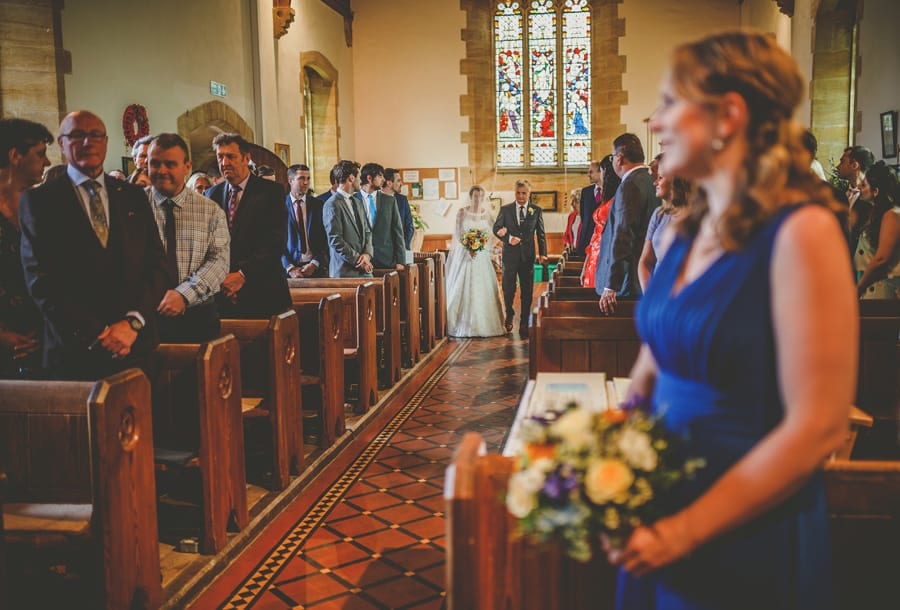 The bride and her father start to walk down the aisle of the church while the wedding guests stand and watch