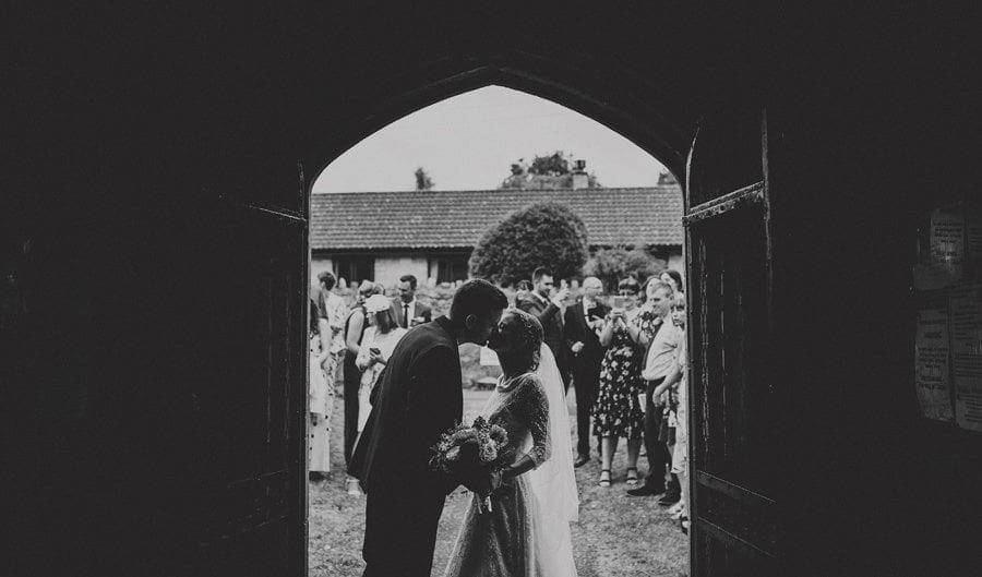 The bride and groom kiss each other at the entrance to the church as the wedding guests stand outside