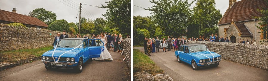 The bride and groom enter a car and drive away from the church as wedding guests stand and watch