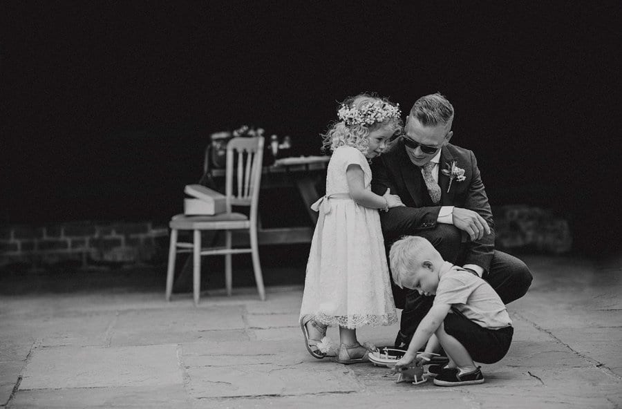 The groom kneels down and listens to his daughter in a courtyard with a little boy in front of them playing with a toy helicopter