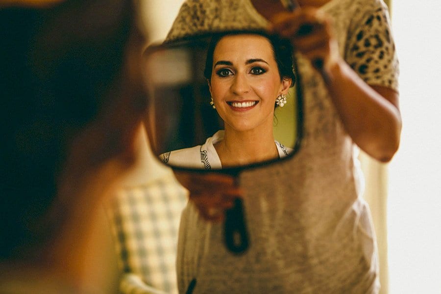 The bride smiles in a mirror held in place by the makeup artist at Wilderness Reserve