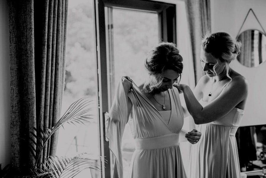 A Bridesmaid helps another bridesmaid to put on their dresses next to a window of the cottage