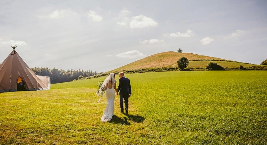 The bride and groom hold hands and walk towards the large tipi in the field at yurt retreat