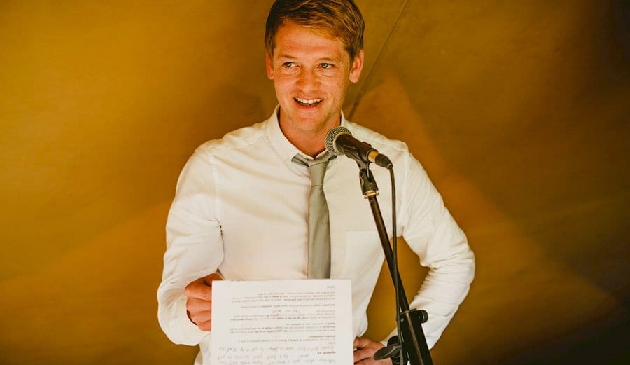 The best man holds a piece of paper in his hand and delivers his speech to the wedding guests in the tipi