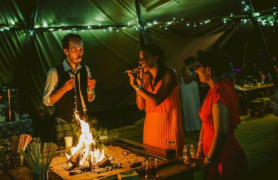 Wedding guests toast marsh mellows over an open lit fire in the tipi