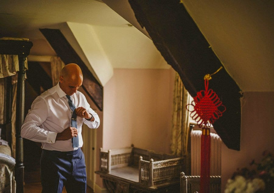 The groom looks down and straightens his tie in a bedroom at Abbey House Gardens