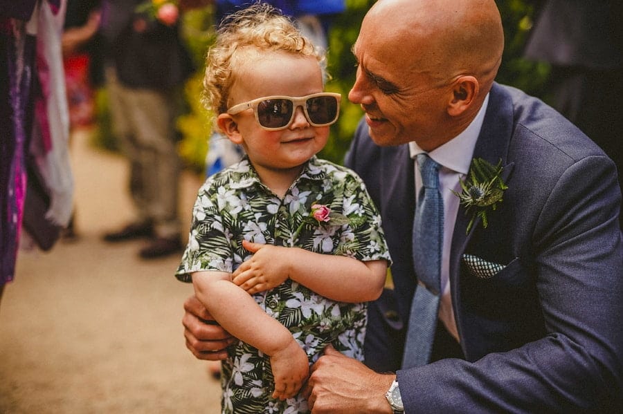The groom smiles at his son wearing sunglasses in the gardens outside Abbey House