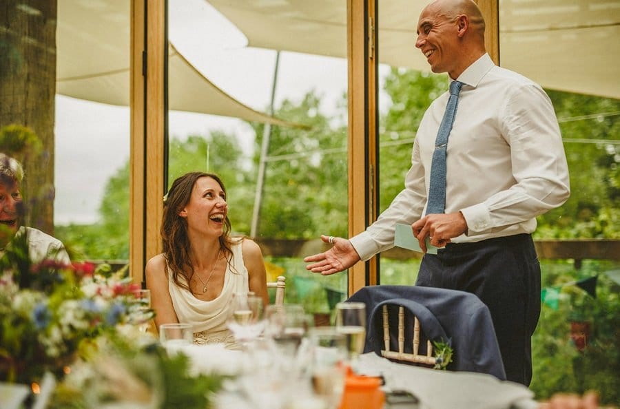 The groom stands up and delivers his speech as the bride looks on and laughs