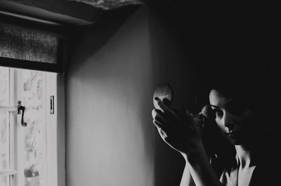 A bridesmaid applies makeup to her face next to a window in one of the bedrooms of the cottage
